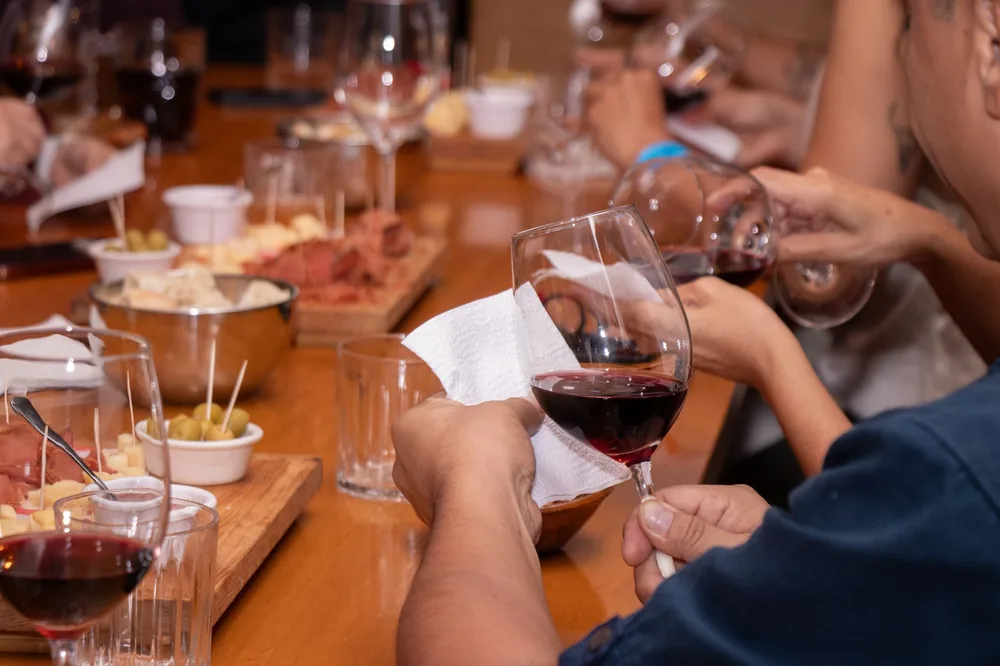 People's hands holding wine glasses at a wine tasting event with snacks on a wooden table