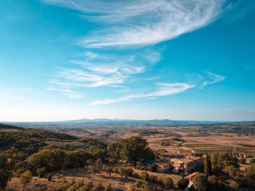 Rural landscape with a farmhouse, rolling hills, lush trees, and a summer sky