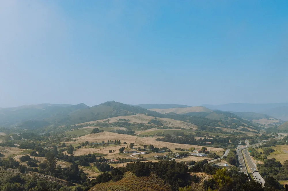 Scenery of hills covered with green trees located near asphalt road under cloudless blue sky