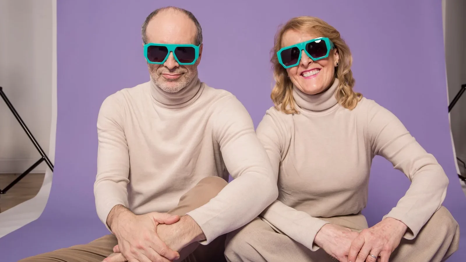 Senior couple wearing matching beige outfits and sunglasses sitting against a purple studio backdrop