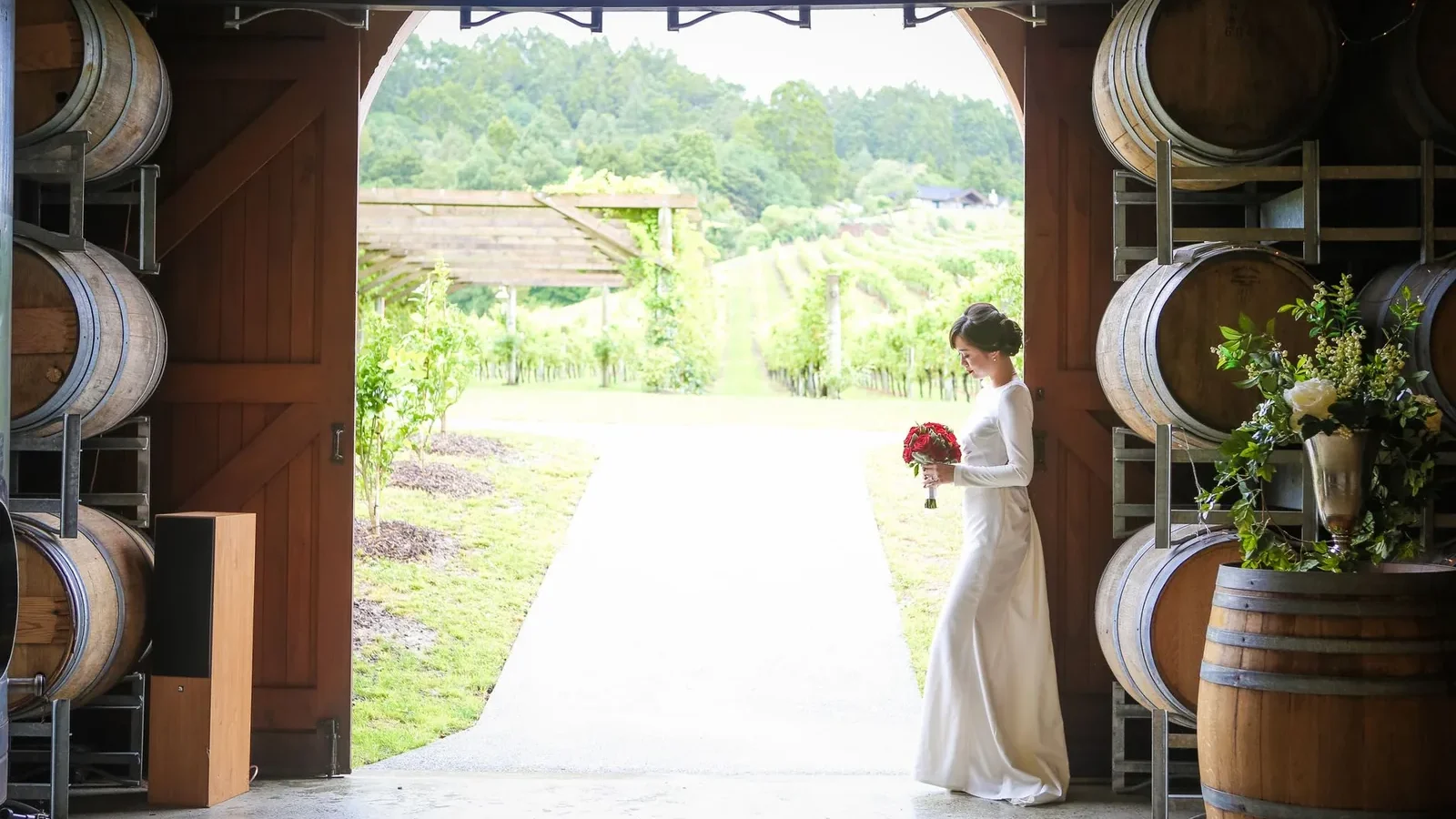 Side view full body of bride in white wedding dress with bouquet of red roses standing in building with wooden barrels in winery near plantation with green plants