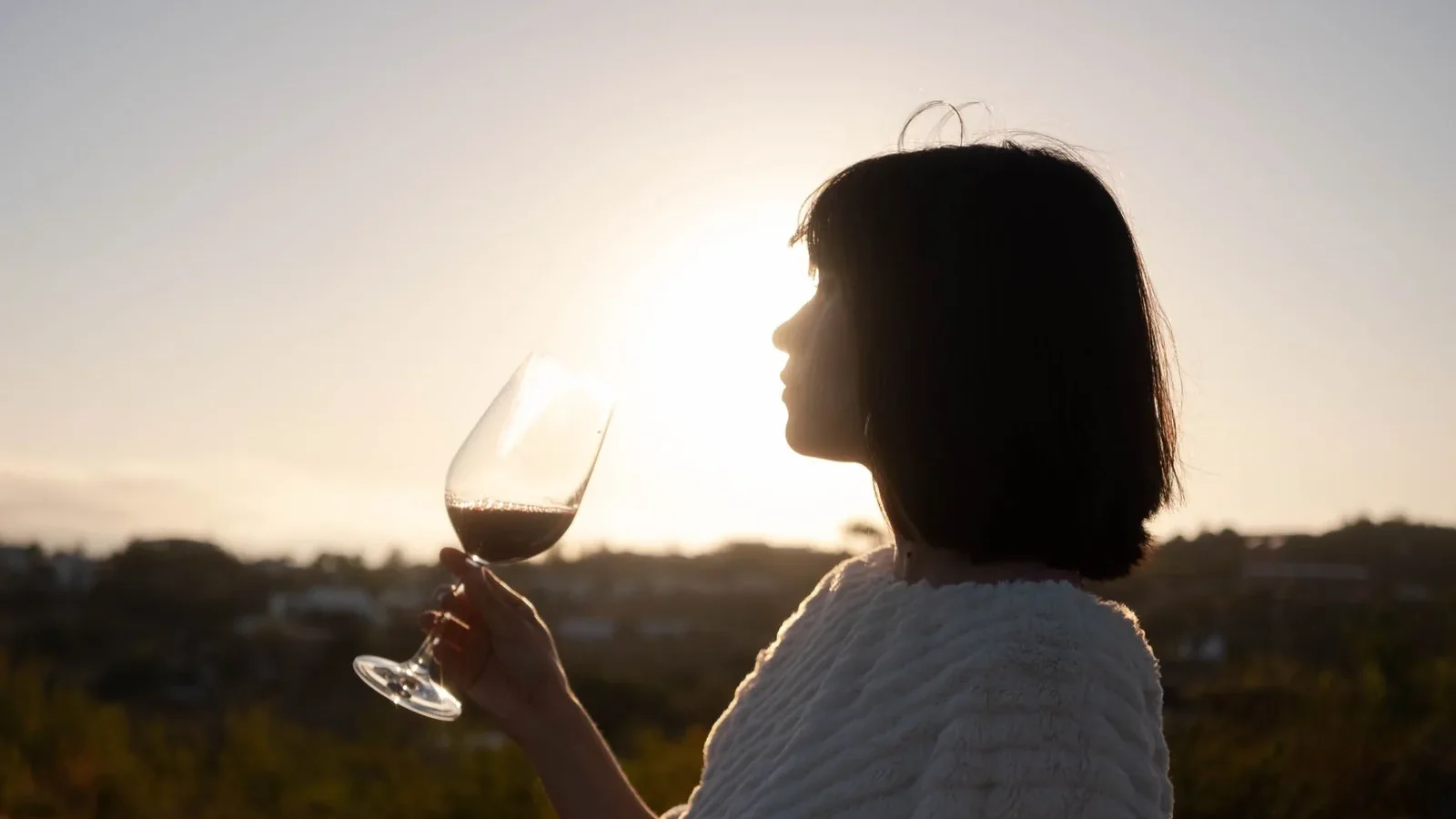 Silhouette of a woman with short hair holding a wine glass, enjoying a serene sunset outdoors