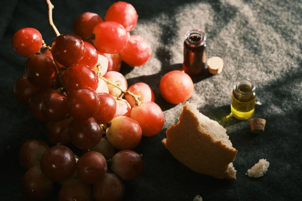 Still life featuring grapes, broken bread, and small bottles on a dark fabric background