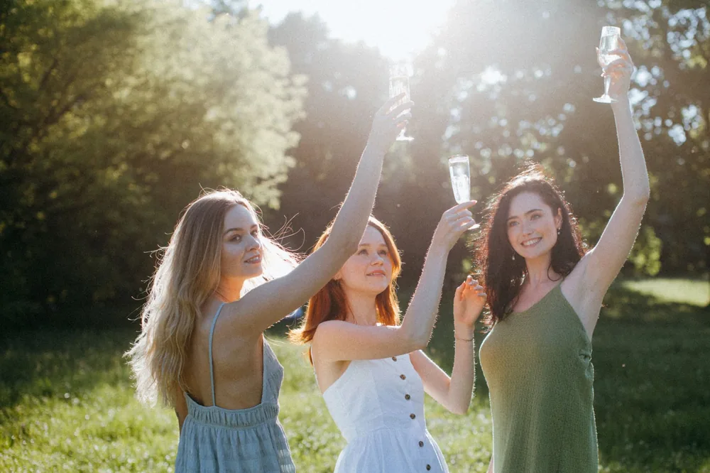 Three women celebrating outdoors with glasses of champagne in summer sunlight, embracing happiness and togetherness