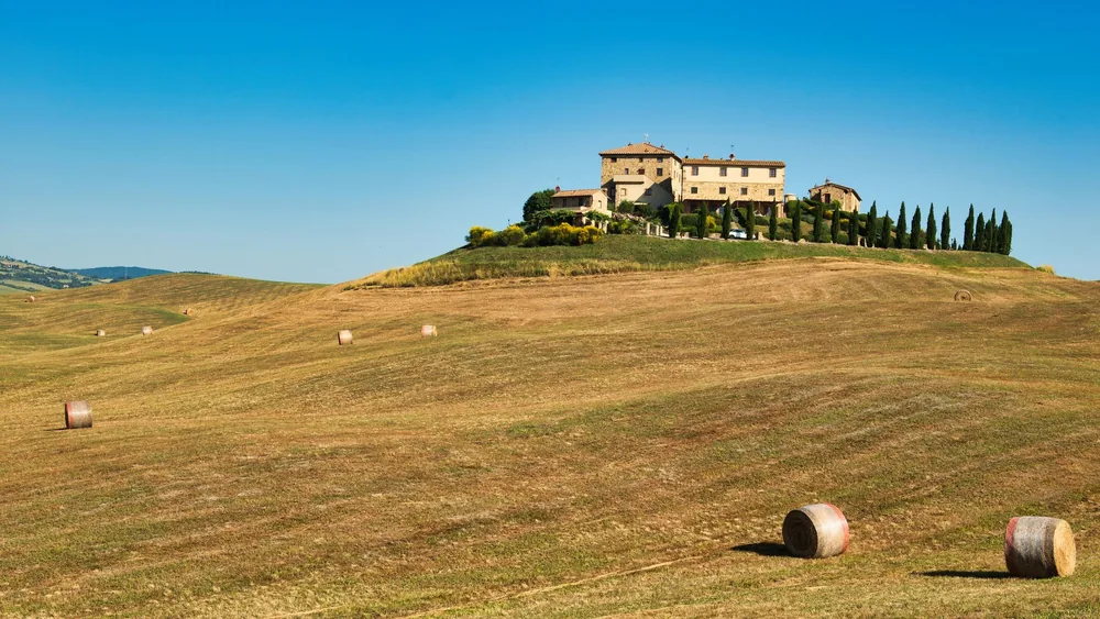 Tuscan farmhouse atop rolling hills surrounded by hay bales under a clear summer sky
