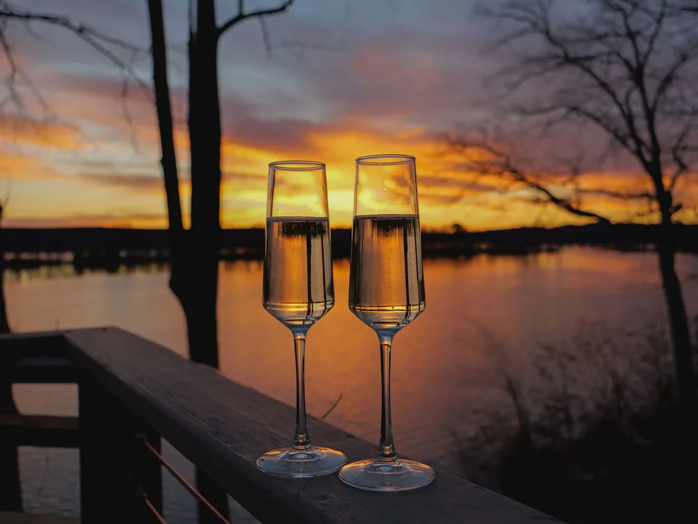 Two champagne glasses at sunset on a balcony overlooking a serene lake