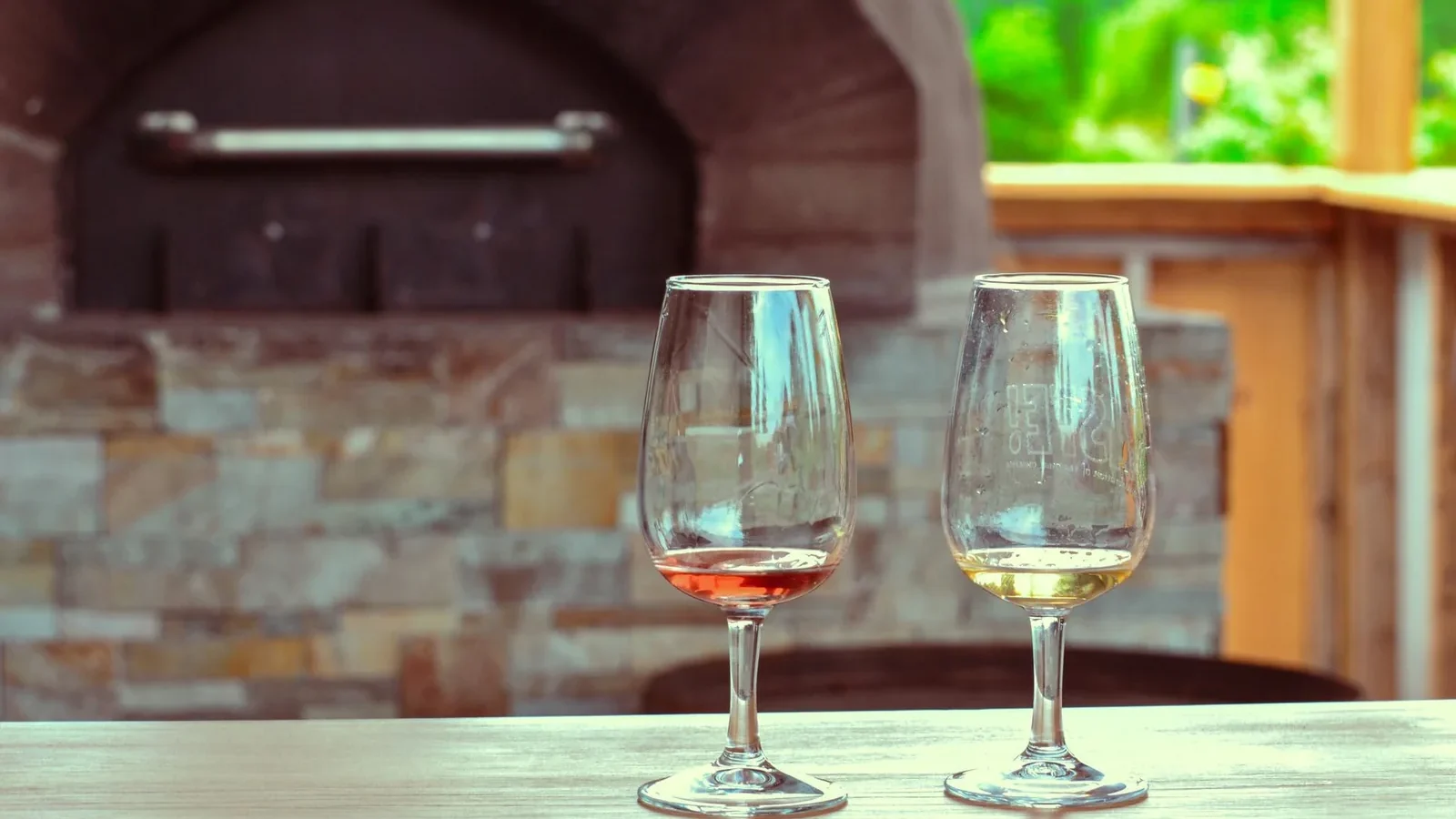 Two empty wine glasses on a table in an outdoor setting with a stone oven in the background