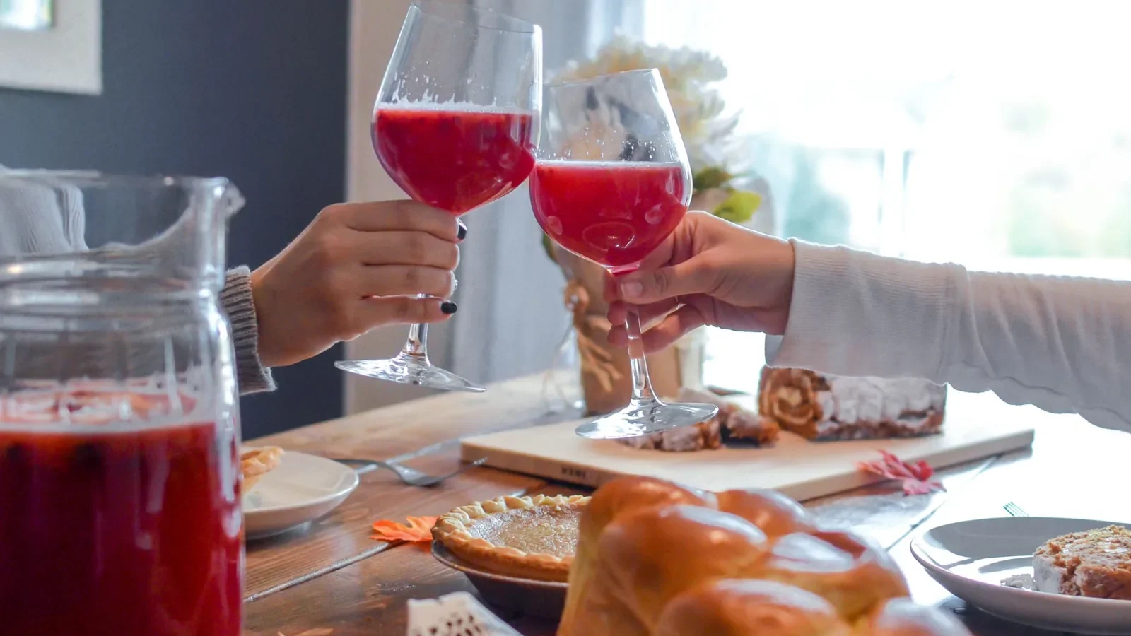 Two people clink glasses with red beverages in a cozy indoor setting surrounded by pastries and pie