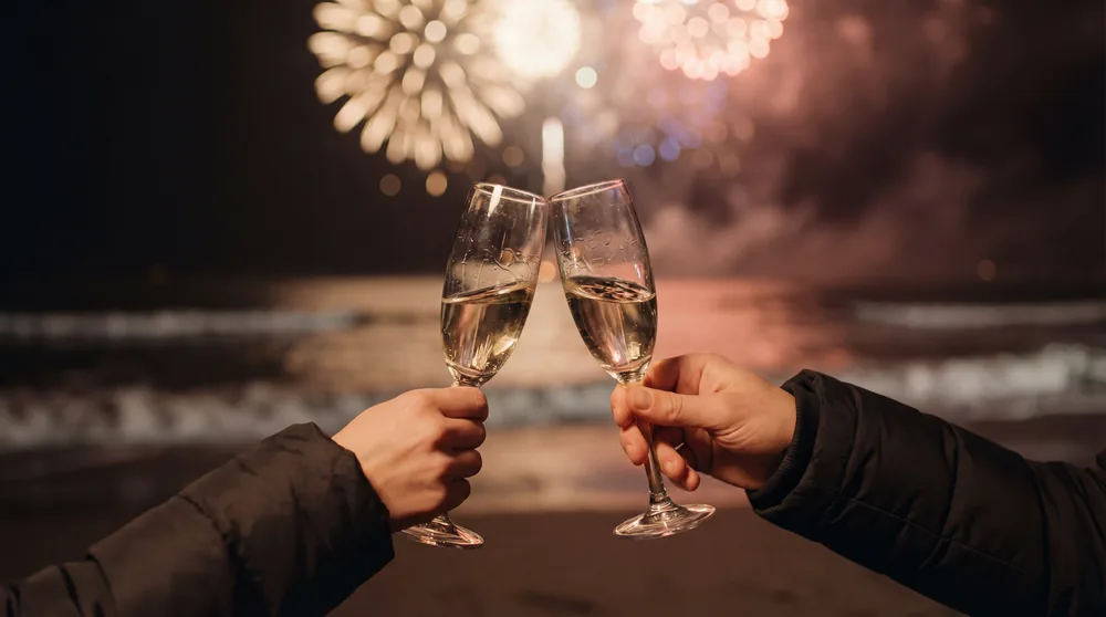 Two people clinking champagne glasses on a beach with fireworks in the background