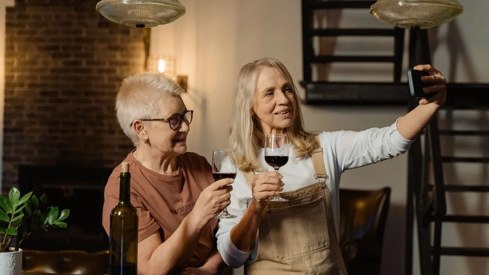 Two senior women enjoying wine indoors, capturing a selfie moment with smiles and camaraderie