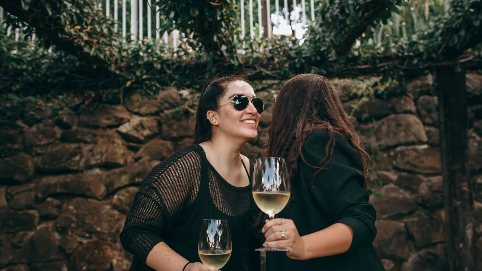 Two women enjoying wine and laughter outdoors against a stone wall backdrop