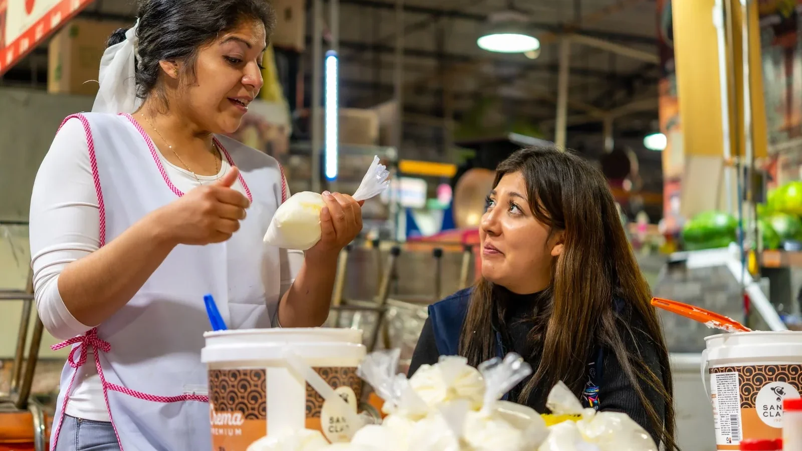Two women interacting at a food market, surrounded by dairy products and produce