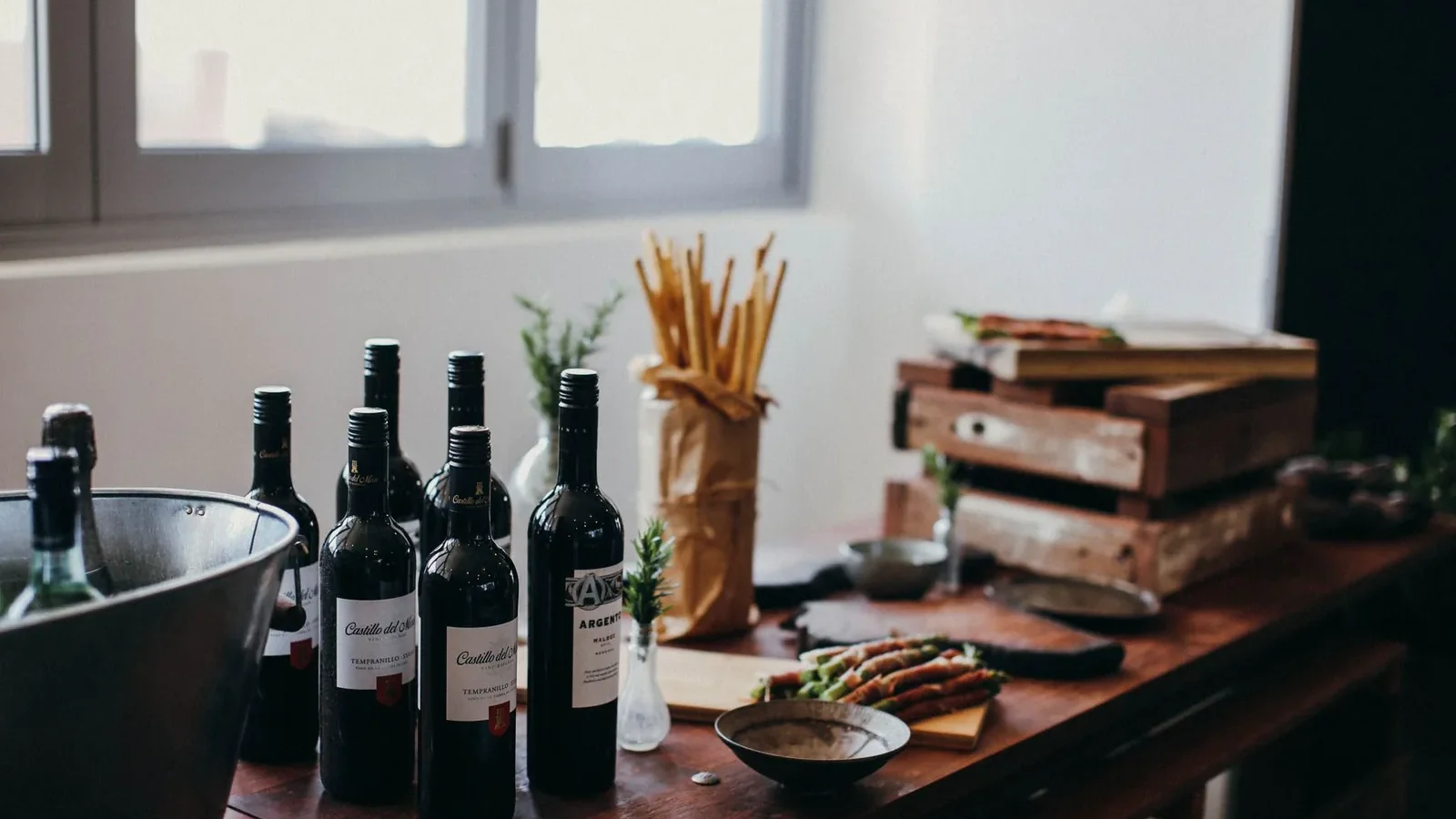 Various bottles of alcoholic drinks placed on table with grissini sticks and wooden boxes near green branches in vase in room