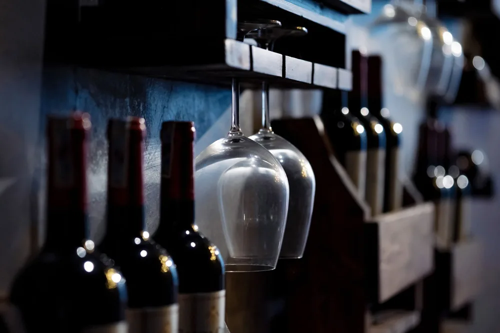 Wine bottles and glasses on wooden shelves in a dimly lit cellar, creating a cozy ambiance