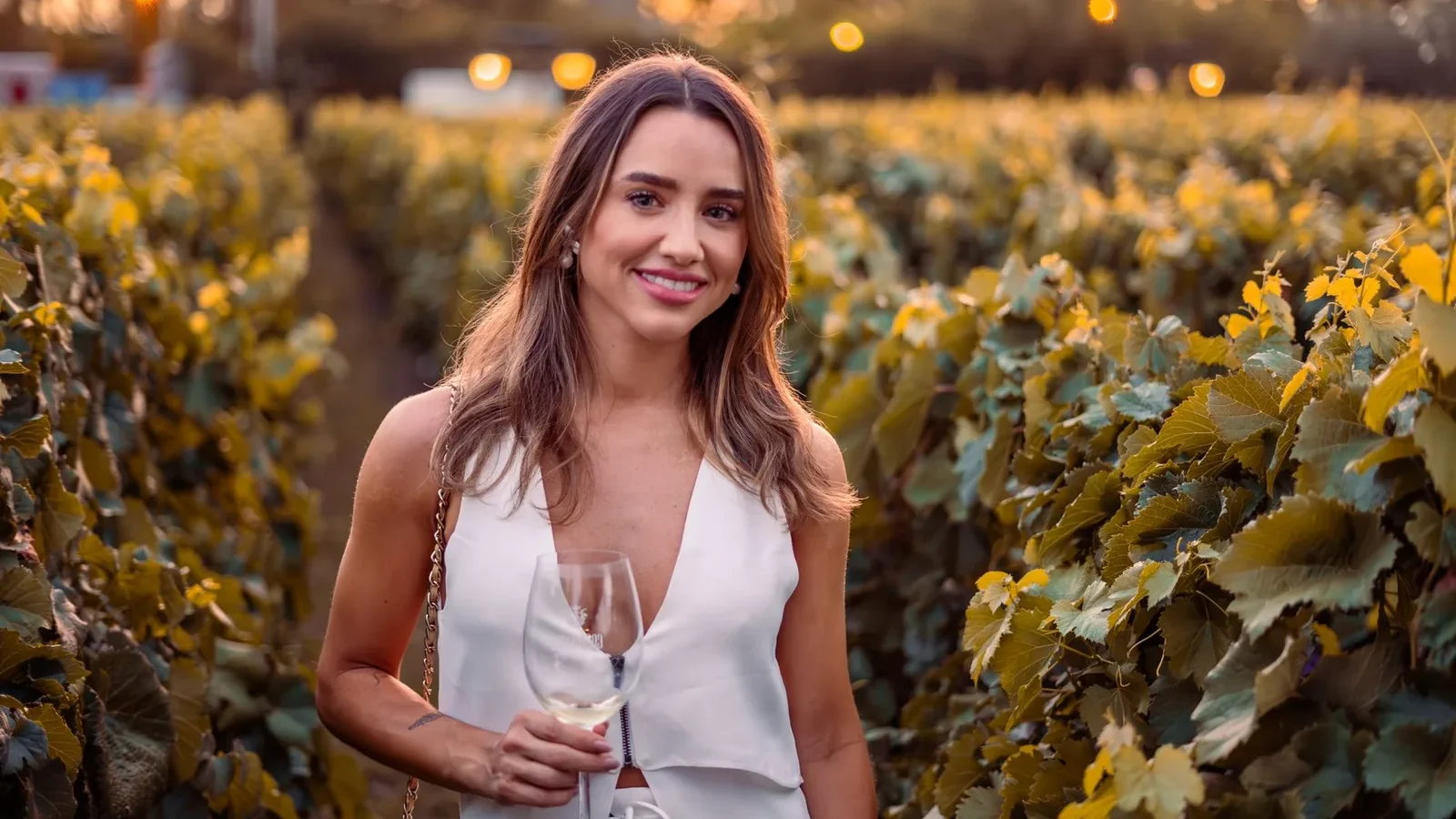 Woman with long brown hair enjoying a drink in a scenic vineyard during sunset